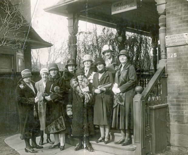Un groupe de femmes habillées à la mode se tiennent devant un immeuble portant une petite enseigne avec la mention Women's College Hospital dans une photo en noir et blanc.