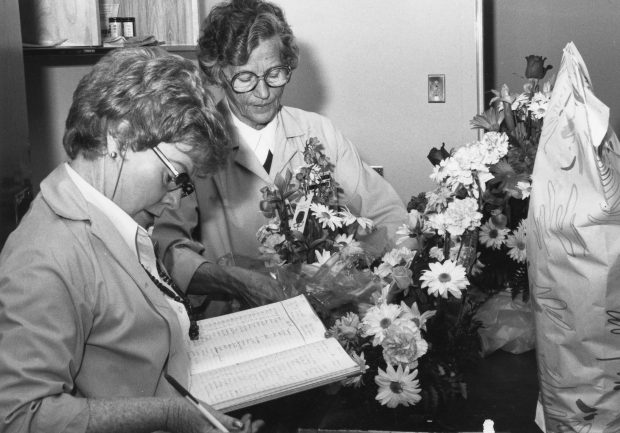 Deux femmes; la première écrit dans un registre, l’autre arrange un bouquet dans une photo en noir et blanc.