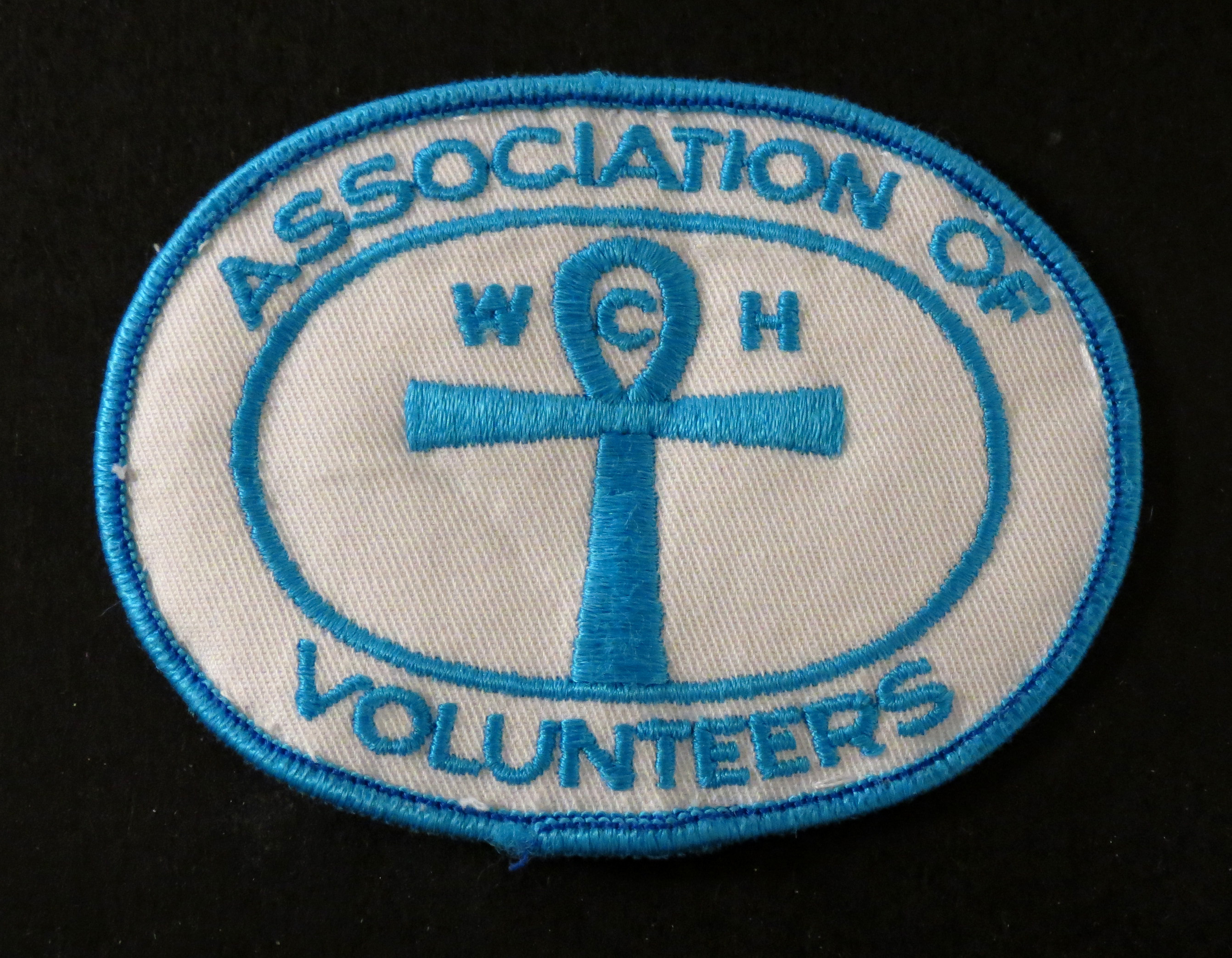 Insigne blanc avec couture bleue. Le texte dit : Association of Volunteers (Association des bénévoles)