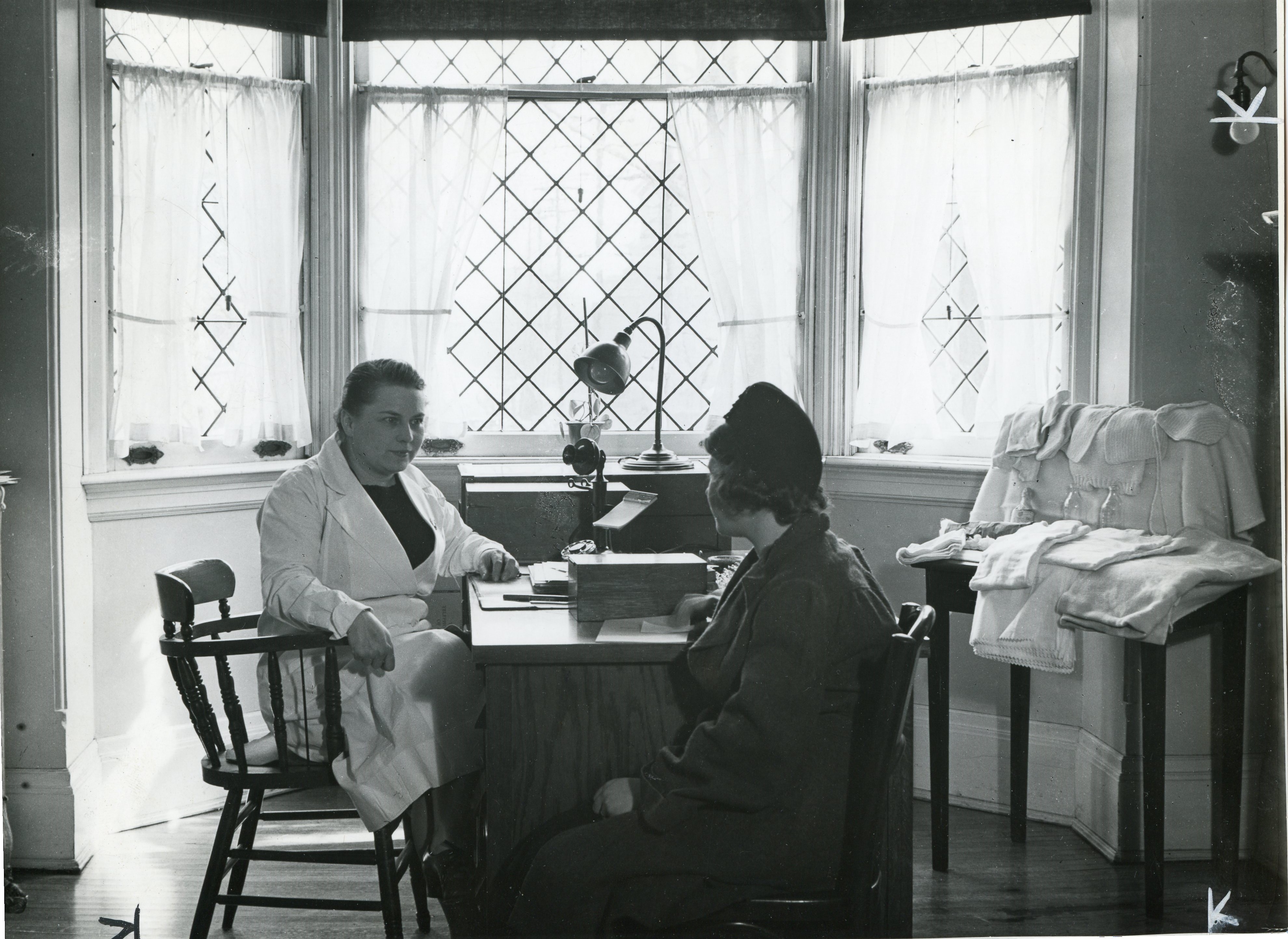 Un professionnel médical en consultation avec une femme à son bureau situé devant une fenêtre en baie dans une photo en noir et blanc. Des linges de bébé et vêtements tricotés sont étalés sur une petite table à droite. 