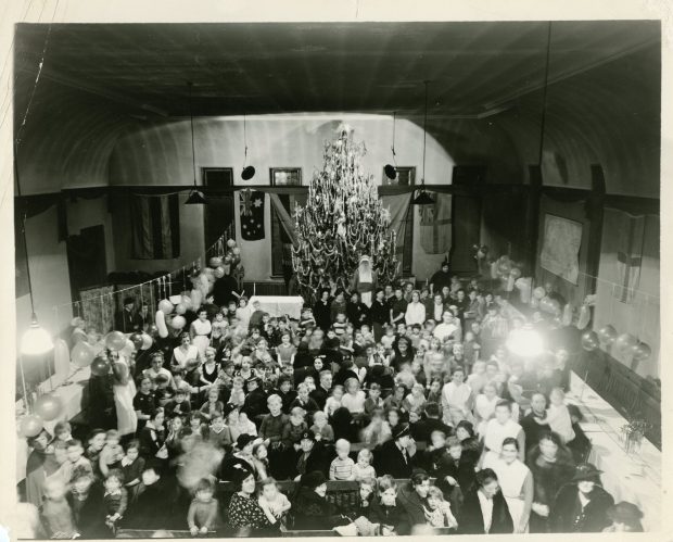 Femmes, enfants et infirmières rassemblés autour de tables longues dans une photo en noir et blanc. Des ballons sont suspendus et le Père Noël se tient près du grand sapin au fond de la salle.