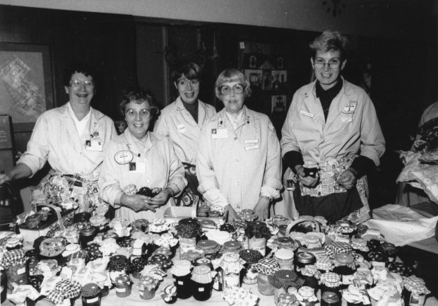 Des femmes en pardessus identiques se tiennent devant une table couverte de fruits en conserve dans une photo en noir et blanc.