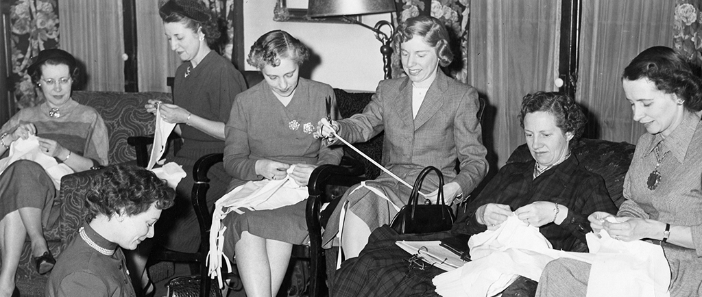 Des femmes travaillent à des œuvres de couture individuelles dans une photo en noir et blanc. Des chaises ont été ajoutées à la pièce pour les accueillir. Une femme est assise à terre dans le coin gauche.