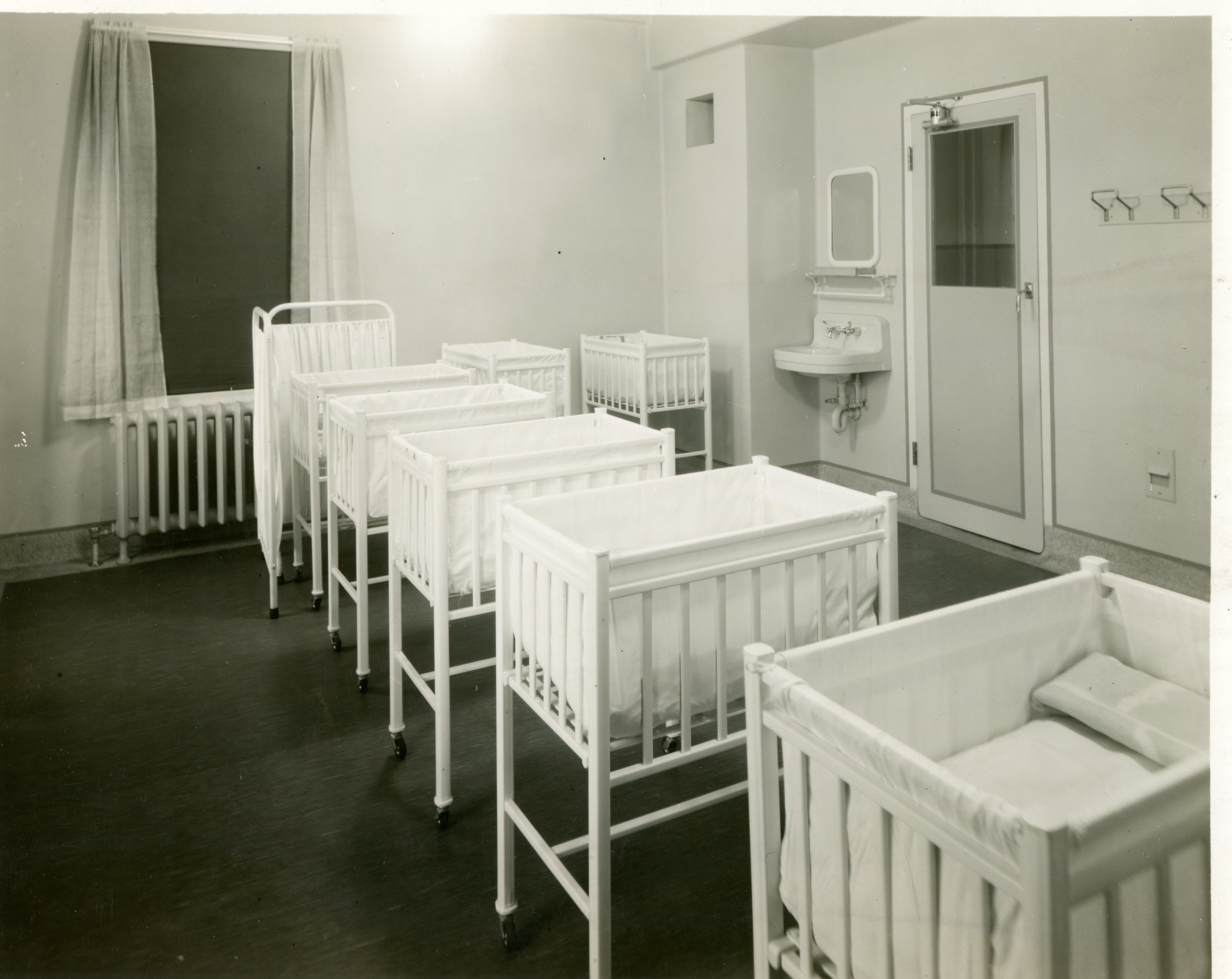 Lits de bébé nombreux dans une pièce claire et ouverte dans un hôpital, avec lavabo et miroir dans le coin près de la porte dans une photo en noir et blanc.