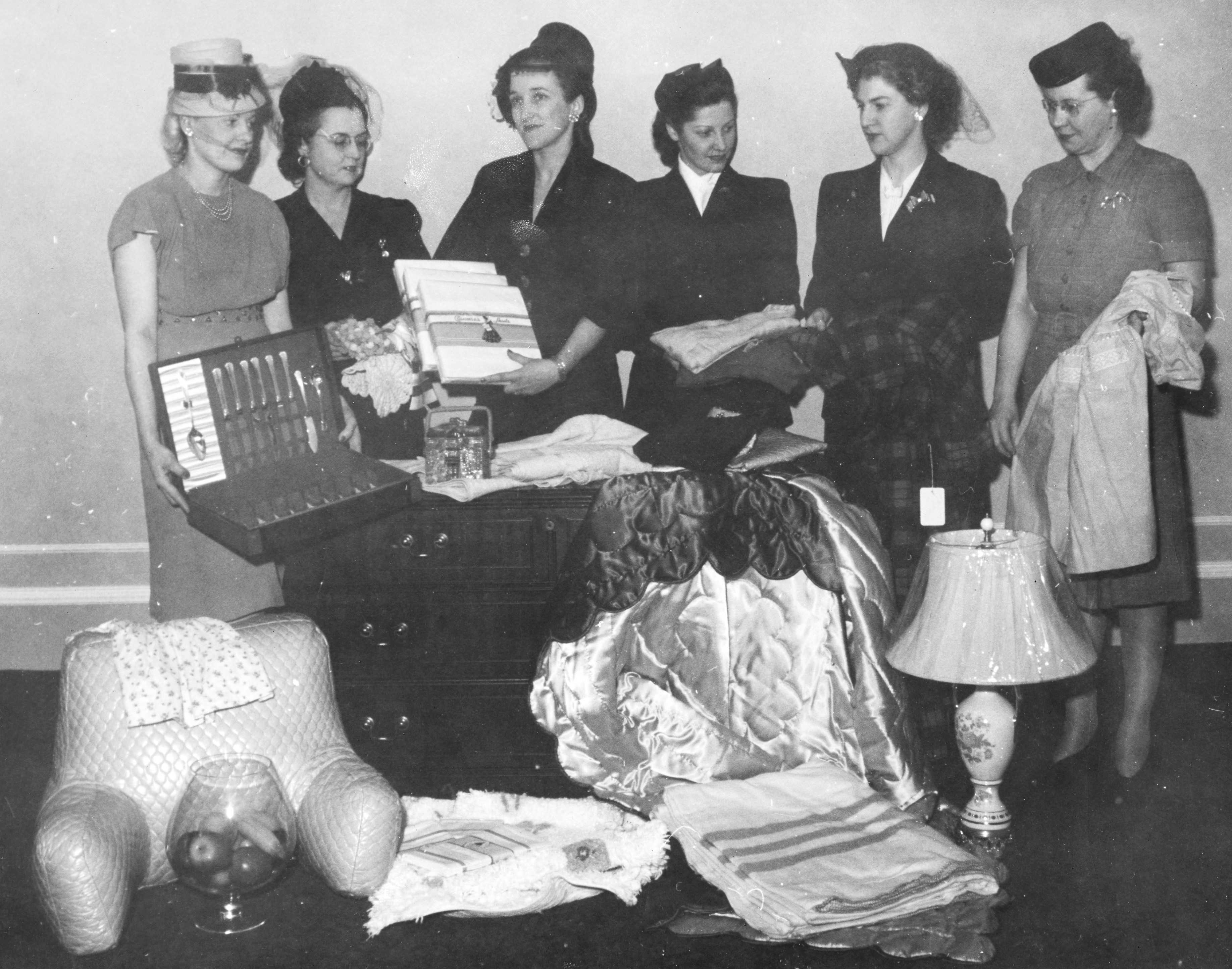 Six femmes en tenue formelle se tiennent derrière un arrangement d’objets ménagers dans une photo en noir et blanc. 