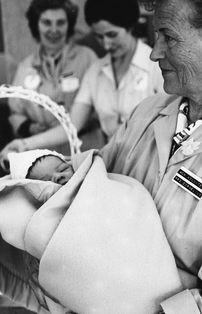 Dans une photo en noir et blanc, une femme en manteau de bénévole tient un bébé endormi enveloppé d’une couverture; deux autres femmes se trouvent en toile de fond. 