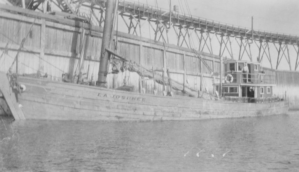 Photographie ancienne et en noir et blanc de La Josuhéee. Le bateau est en bois, la voile est baissée et on peut lire aisément le nom du bateau. À l'arrière, le capitaine Joseph Desgagnés se tient fièrement à l'extérieur de sa cabine. Au deuxième plan, on observe un quai de bois. 