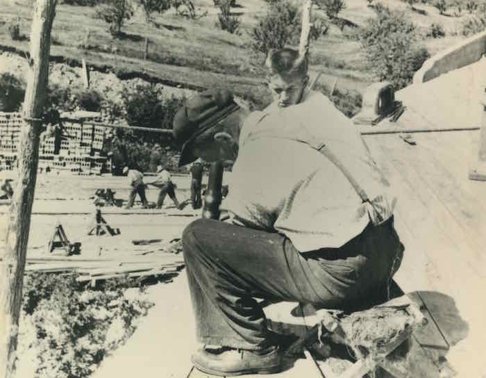 Photographie en noir et blanc. Deux personnes apparaissent au premier plan. Ils semblent assis sur la coque d'un navire. Un homme est assis avec un maillet à la main. Un jeune homme, placé juste derrière, l'observe attentivement. Au second plan, on devine un chantier : matériaux de construction et hommes au travail complètent la scène.