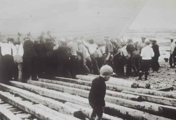 Photo ancienne en noir et blanc. Au premier blanc un jeune enfant marche seul sur des planches de bois. À l'arrière, une vingtaine d'hommes poussent une goélette vers le fleuve.