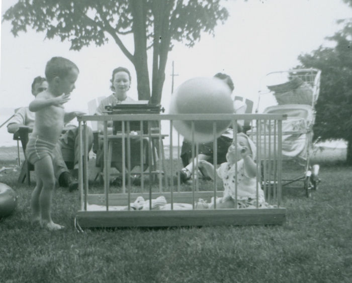 Sur cette photo en noir et blanc prise à l’extérieur apparaît au premier plan un bébé dans un parc. Il joue avec un ballon. À sa gauche, sur le gazon, un jeune garçon se tient debout. À l’arrière, trois adultes sont assis. Au centre, on observe une femme qui travaille à la machine à écrire. La dactylo est déposée sur une petite table dans le gazon. 