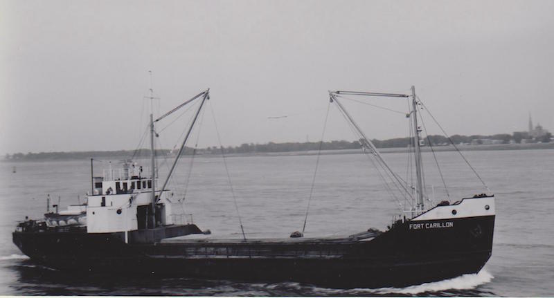 Photographie en noir et blanc. On observe le caboteur Fort Carillon dans toute sa longueur, vue de côté. La coque du bateau est noire et son nom est inscrit en lettres blanches à la proue. Il a deux mâts de charge et flotte sur l’eau. Derrière, au loin, on peut voir un petit hameau et le clocher d’une église. 