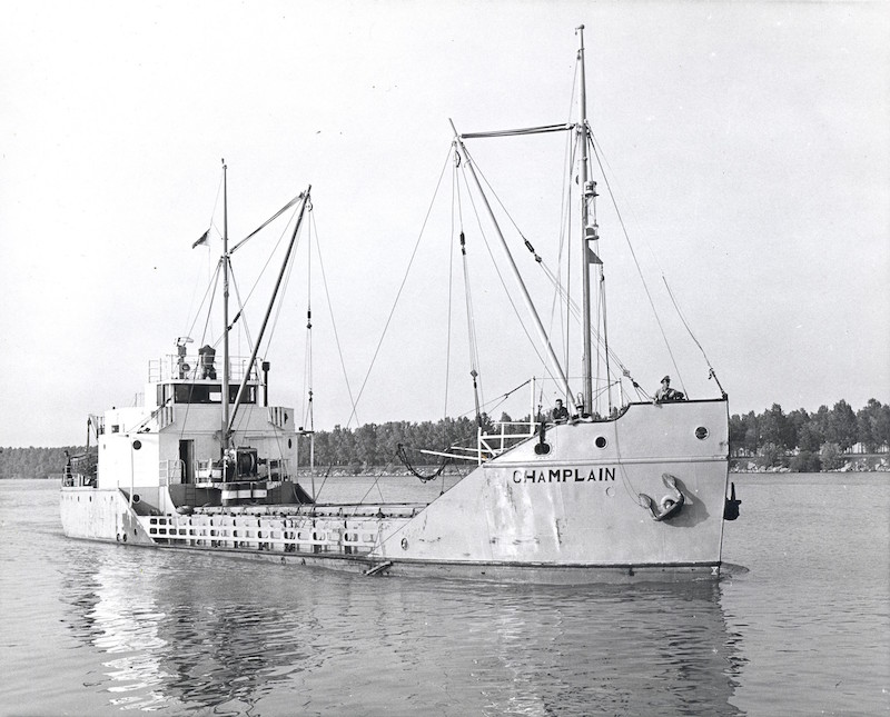 Photographie en noir et blanc. On observe le caboteur Champlain dans toute sa longueur, vue de devant. Le bateau est pâle et son nom est inscrit en lettre noire à la proue. Il a deux mâts de charge et ne semble transporter aucune cargaison. Trois hommes se tiennent au-devant du bateau qui flotte sur l’eau.