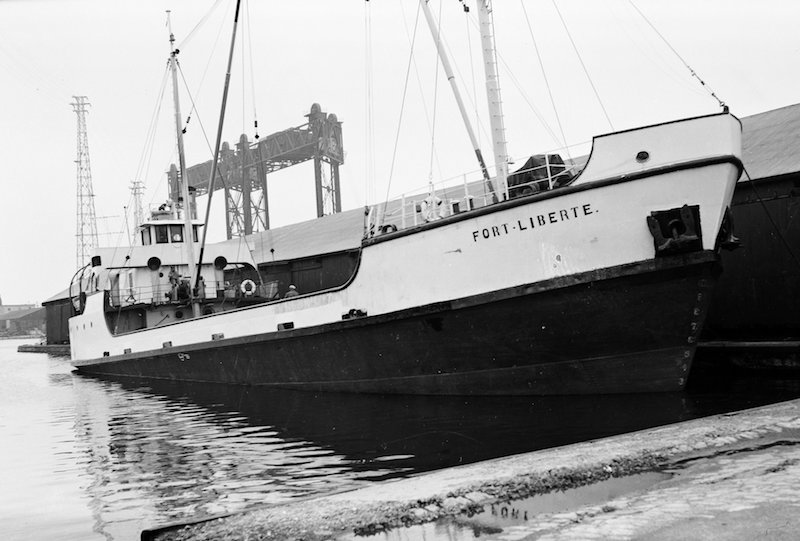 Photographie en noir et blanc. On observe le caboteur Fort Liberté dans toute sa longueur, vue de devant. La coque du bateau est noire et blanche et son nom est inscrit en lettres noires à la proue. Il a deux mâts de charge et ne semble transporter aucune cargaison. Derrière, on observe les structures d’une écluse. 