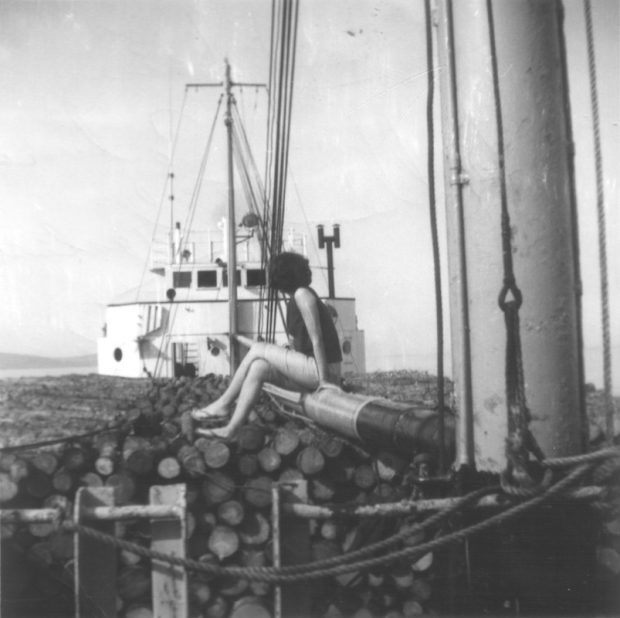 Photographie en noir et blanc prise sur le pont du navire, à la poupe. Le navire est rempli de cordes de bois, sur lesquelles est assise une femme de dos. Devant elle, un mât de charge et la cabine blanche du navire. 