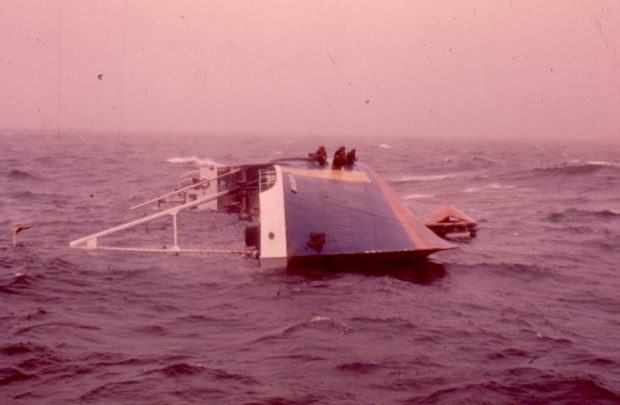Photographie couleur montrant l’Aigle d’Océan à demi submergé. Le photographe se tient devant la coque du bateau. Le navire est renversé sur son flanc et ses mâts sont plongés dans l’eau. Sur la coque bleue et jaune, des membres d’équipage attendent les secours. 