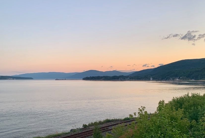 Vue du fleuve Saint-Laurent au coucher de soleil. Au premier plan, un rail de chemin de fer et des arbustes. Au second plan, une étendue d’eau aux reflets rosés. Quelques nuages parsèment le ciel bleu et rose. Au loin, des montagnes. 