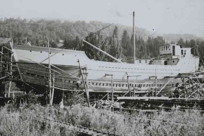 Cette photographie en noir et blanc montre une goélette en construction. Le bateau semble presque achevé. Des échafaudages de bois se dressent de part et d'autre de la charpente. Quelques hommes - très petits sur la photo - s'affairent sur le bateau. Devant le bateau, un homme se tient dans les herbes hautes et observe la scène.