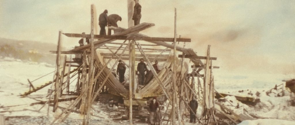 Vue d'une goélette en construction. On y voit la charpente du bateau entourée par des échafaudages. Des hommes en habits de travail se tiennent dans la cale du bateau et sur un échafaudage. Devant, se tiennent une femme, un enfant et un autre homme. La photo a été prise en hiver. On a coloré l'image, ce qui lui donne une teinte sépia.