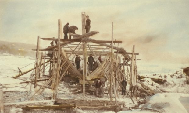 Vue d'une goélette en construction. On y voit la charpente du bateau entourée par des échafaudages. Des hommes en habits de travail se tiennent dans la cale du bateau et sur un échafaudage. Devant, se tiennent une femme, un enfant et un autre homme. La photo a été prise en hiver. On a coloré l'image, ce qui lui donne une teinte sépia.