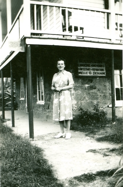 Photographie en noir et blanc. Une femme portant une robe pâle à motifs se tient sous la galerie d'une maison. À la base, des murs de pierre, au-dessus, des murs de bois. Sur le mur de pierre est apposée une affiche sur laquelle on peut notamment lire Melle G.Desgagne.