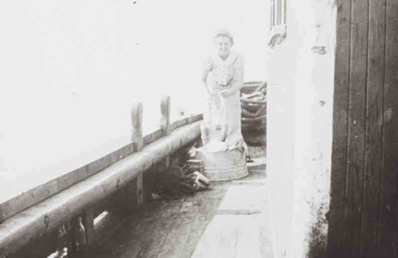 Photographie en noir et blanc. Une jeune femme souriante, les cheveux attachés se tient debout sur le pont d’un navire. Elle semble faire une corvée de vaisselle dans des seaux de métal. Quelques cordages sont enroulés devant et derrière elle. 