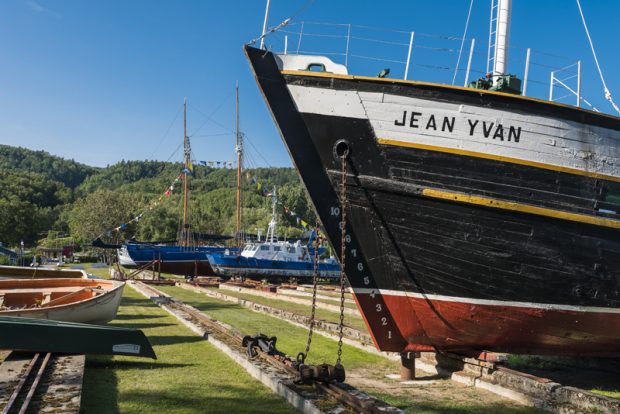 Photographie du terrain du Musée maritime de Charlevoix. Au premier plan, on aperçoit une partie de la coque de la goélette Jean Yvan, peinte en blanc, noir, jaune et rouge. Des rails forment des lignes droites au bout duquel on observe d’autres bateaux parés de drapeaux. Sur la gauche, des chaloupes reposent sur le sol. 