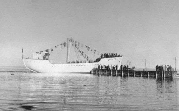 Vue en noir et blanc d’une goélette sur le fleuve Saint-Laurent. Au premier plan, on observe une étendue d’eau. Sur la droite de l’image, un quai s’avance dans le fleuve. Plusieurs personnes se tiennent sur le quai et observe le majestueux bateau blanc décoré de plusieurs pavillons maritimes. D’autres personnes sont à bord de la goélette. 