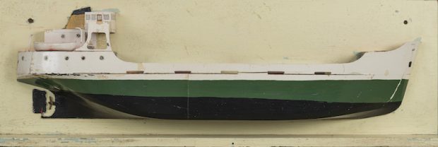 Cette photo en couleur présente une maquette du caboteur Mont St-Martin. Il s'agit d'une coupe longitudinale. La coque du bateau a une bande noire et une bande verte. La partie supérieure et les cabines sont peintes en blanc. À l'arrière, on aperçoit l'hélice du bateau.