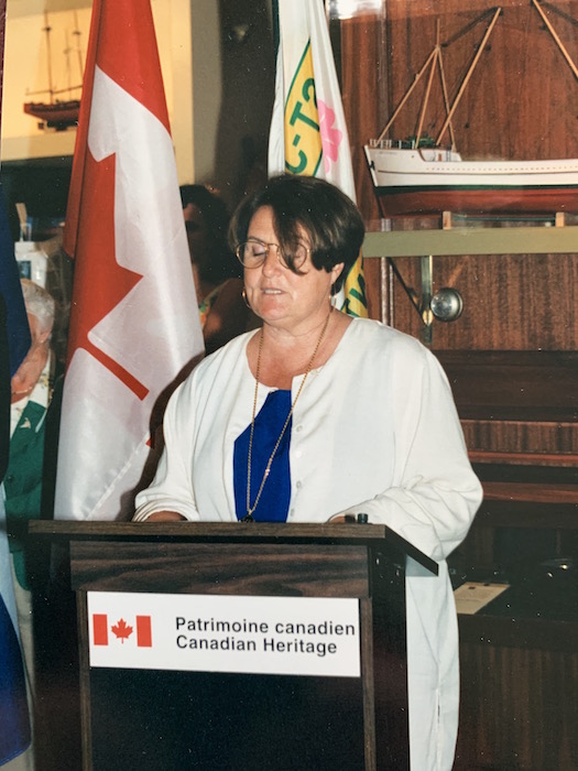 Une femme se tient debout devant un lutrin à l’effigie de Patrimoine Canada. Elle livre un discours. Derrière elle, deux drapeaux et une maquette de bateau. 