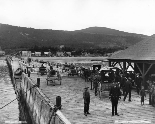 Vue du quai des Éboulements (Saint-Joseph-de-la-Rive), vers 1890. La photo a été prise d’un navire amarré au bout du quai, on voit donc la rive à l’horizon et quelques maisons. Le quai est en bois. Au premier plan, on aperçoit un homme en uniforme. Près de lui se tiennent quelques enfants et d’autres hommes. Au second plan, on y observe des calèches.