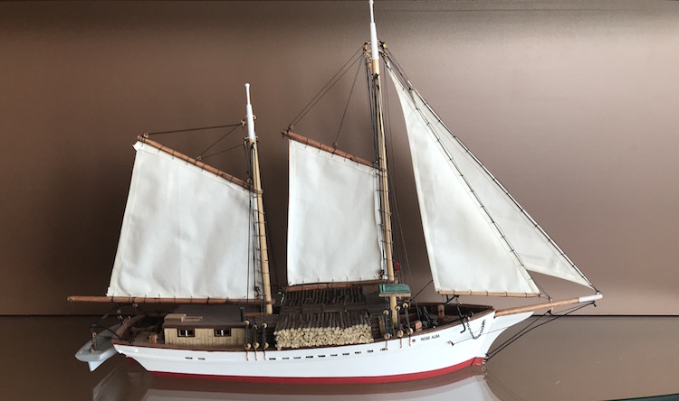 Maquette de la goélette à voiles Rose-Alba. La cale du bateau est blanche, mais a une petite bande rouge est peinte à sa base. Le bateau est garni de cordes de bois miniatures et d'une petite chaloupe de sauvetage à sa poupe. Quatre voiles blanches ornent la maquette.