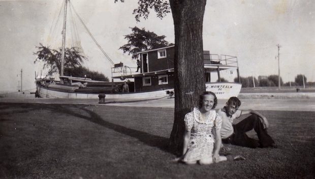 Photographie en noir et blanc. Au premier plan, une jeune femme et un jeune homme sont assis dans l'herbe, accotés sur un arbre. À l'arrière, on observe un bateau de bois sur lequel se dressent trois personnes qui sont difficiles à voir. Le bateau semble flotter sur un canal étroit. 
