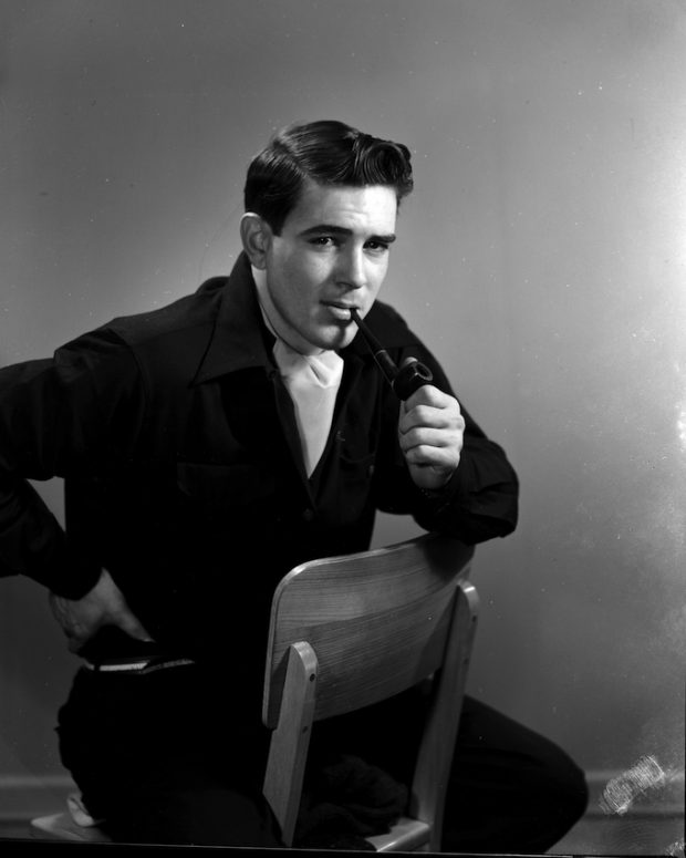 Portrait en noir et blanc d'un jeune homme. Il est assis sur une chaise de bois dont le dossier fait face à son ventre. Le jeune homme est vêtu de noir, porte un foulard blanc noué et a une pipe à la bouche.