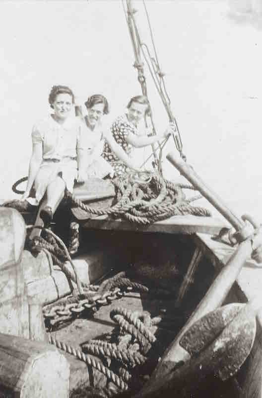 Photographie en noir et blanc montrant trois jeunes femmes se tenant à la proue d'un bateau. À l'avant-plan, on aperçoit des bûches de bois, des cordages et une ancre.