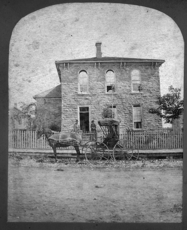 Photographie en noir et blanc d’une maison en pierre avec une voiture hippomobile au premier plan.