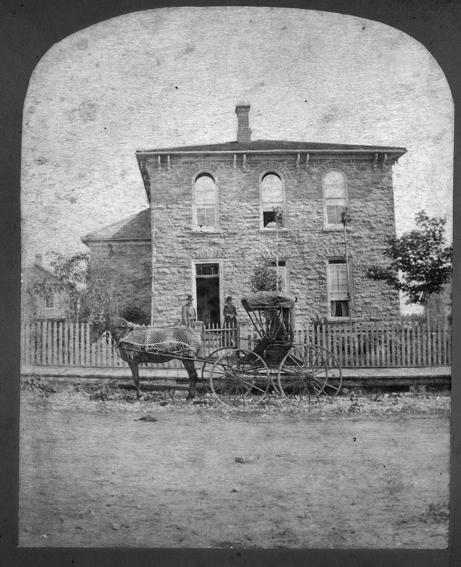 Photographie en noir et blanc d’une maison en pierre avec une voiture hippomobile au premier plan.