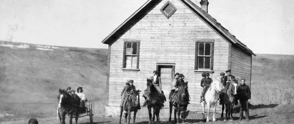 Cinq chevaux alignés devant une école, deux enfants sur chaque cheval. Un cheval et un chariot avec trois enfants et un chien.