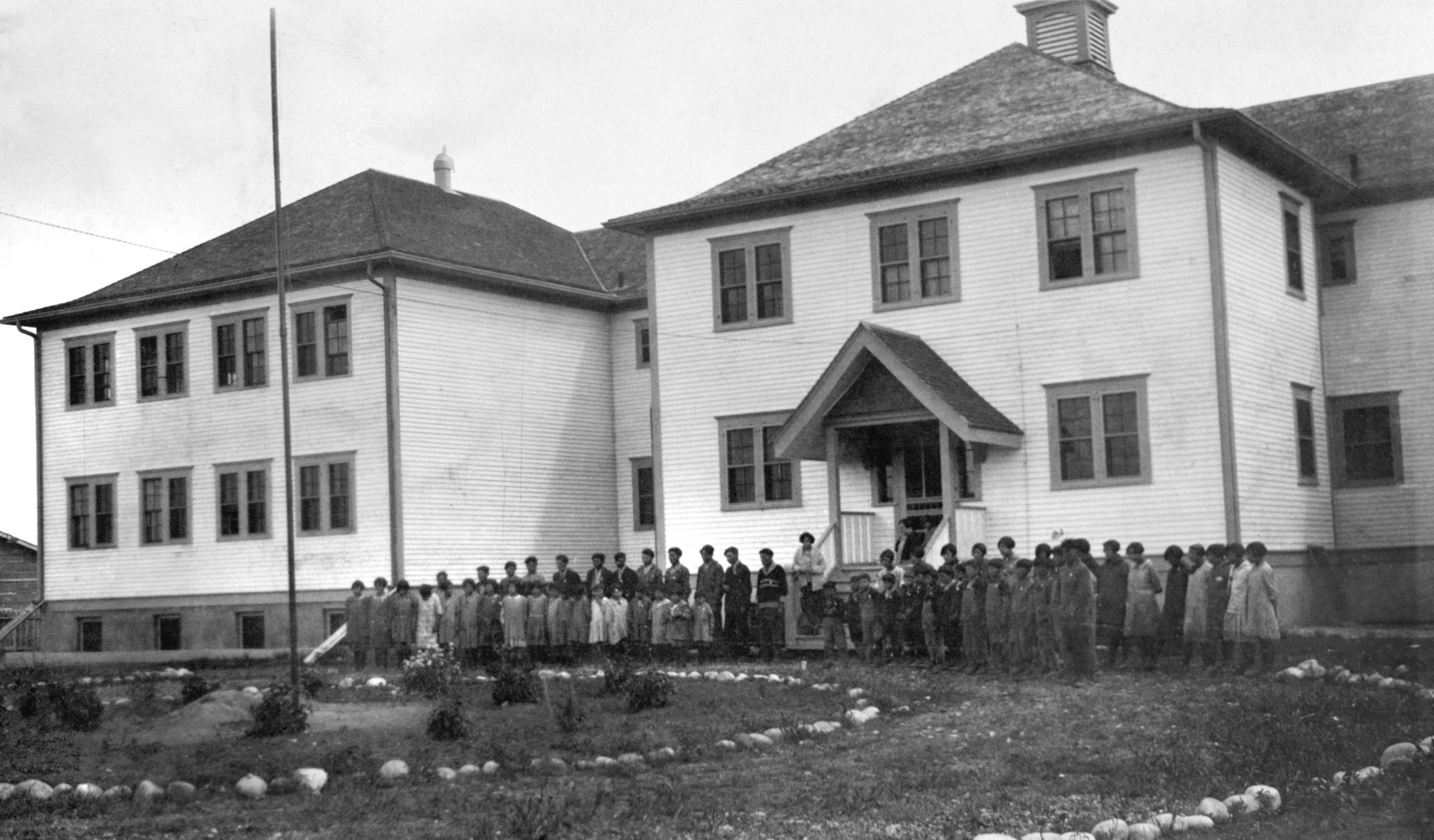 Grand pensionnat à deux étages, enfants et personnel enseignant devant l’école, grand terrain sur lequel des pierres sont aménagées en forme de cercle, et mât de drapeau.