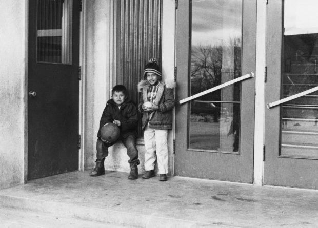 Deux garçons tenant des balles, vêtus de vêtements d'hiver, bottes, manteaux, tuque, pas de gants, debout devant les portes d'entrée.