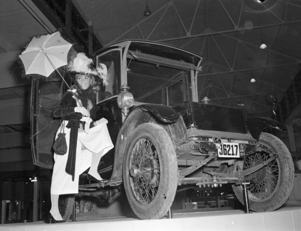 Photo noir et blanc d'un mannequin dans une robe du début du 20e siècle posant dans le cadre de la portière d'une voiture électrique ancienne.
