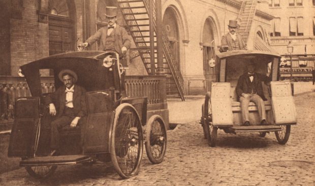 Deux taxis électriques ouverts à l’avant, garés l'un à côté de l'autre dans une scène de rue. Chacun des véhicules transporte un passager et un chauffeur en uniforme portant un chapeau haut de forme.