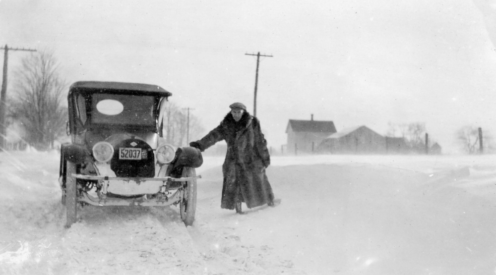 Photo noir et blanc d'un homme en manteau de fourrure appuyé contre une automobile dans la neige profonde. Des bâtiments agricoles et des champs sont visibles à l'arrière-plan.