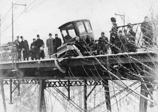 Photo noir et blanc d'une voiture électrique en équilibre sur la rambarde d’un pont par temps de neige. La voiture est entourée de policiers et de badauds.