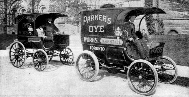 Deux véhicules à moteur de forme rectangulaire et ressemblant à des calèches sont garés devant un bâtiment en pierre. Les conducteurs se détendent sur leur siège. Une inscription sur chacun des véhicules indique : PARKER'S DYE WORKS.