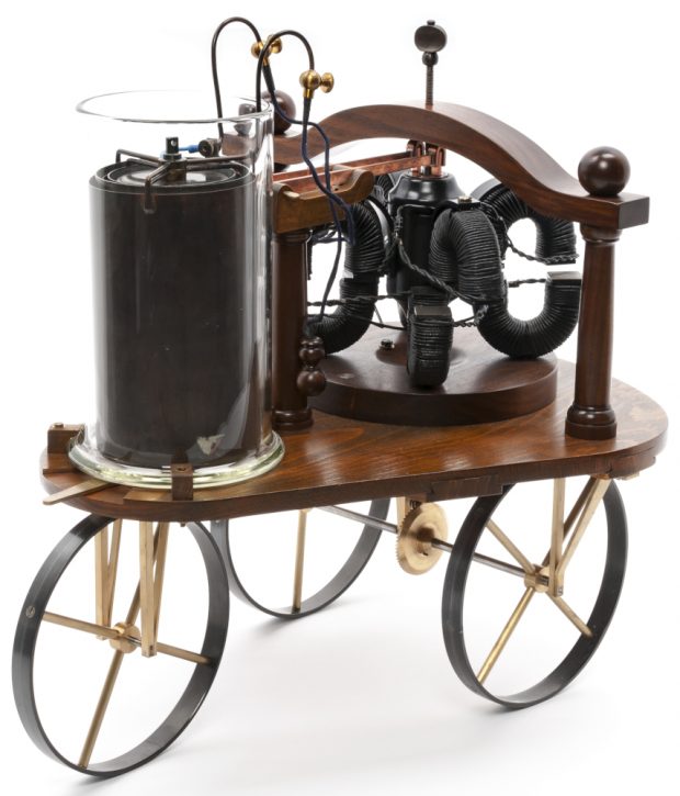 Un véhicule électrique en bois à trois roues, de la taille d'un jouet, avec un moteur exposé et une batterie en verre.