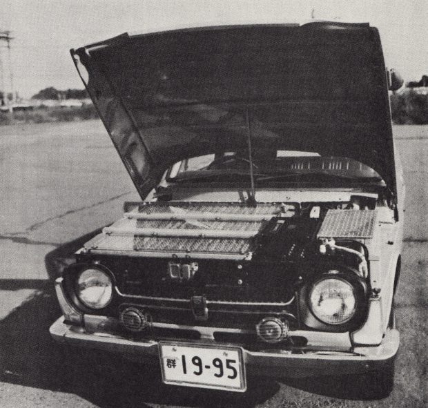 Photo noir et blanc d'une voiture avec une plaque d'immatriculation japonaise et son capot ouvert. Son compartiment moteur est rempli de centaines de cellules de batteries rectangulaires.