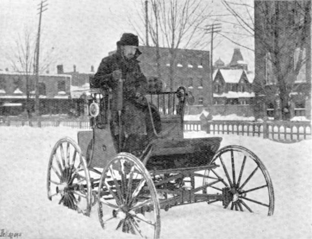 Photo noir et blanc d'un homme moustachu conduisant une voiture électrique à siège élevé dans la neige profonde.