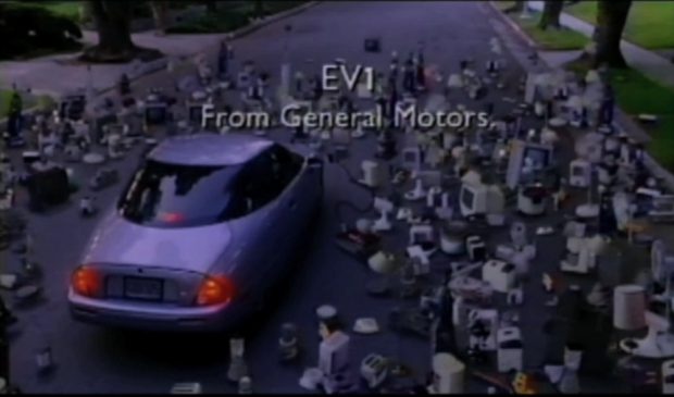 Vue arrière d'une voiture électrique garée au milieu de la route, entourée de centaines d'appareils électriques. Texte : L’EV1 / de General Motors