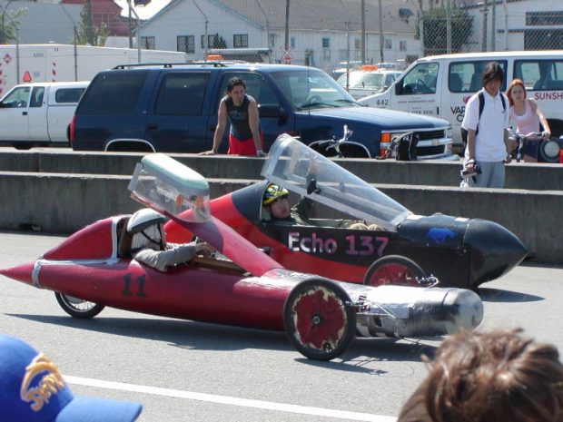 Photo couleur de deux adolescents dans des voitures de course électriques profilées « artisanales » garées au bord d'une piste de course.