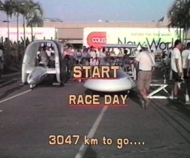 Plusieurs véhicules aux formes insolites sont garés sur la ligne de départ d’une piste de course remplie de monde, entourés de membres de l'équipe et de techniciens. Texte superposé : DÉPART / JOUR DE COURSE / 3 047 km à parcourir...